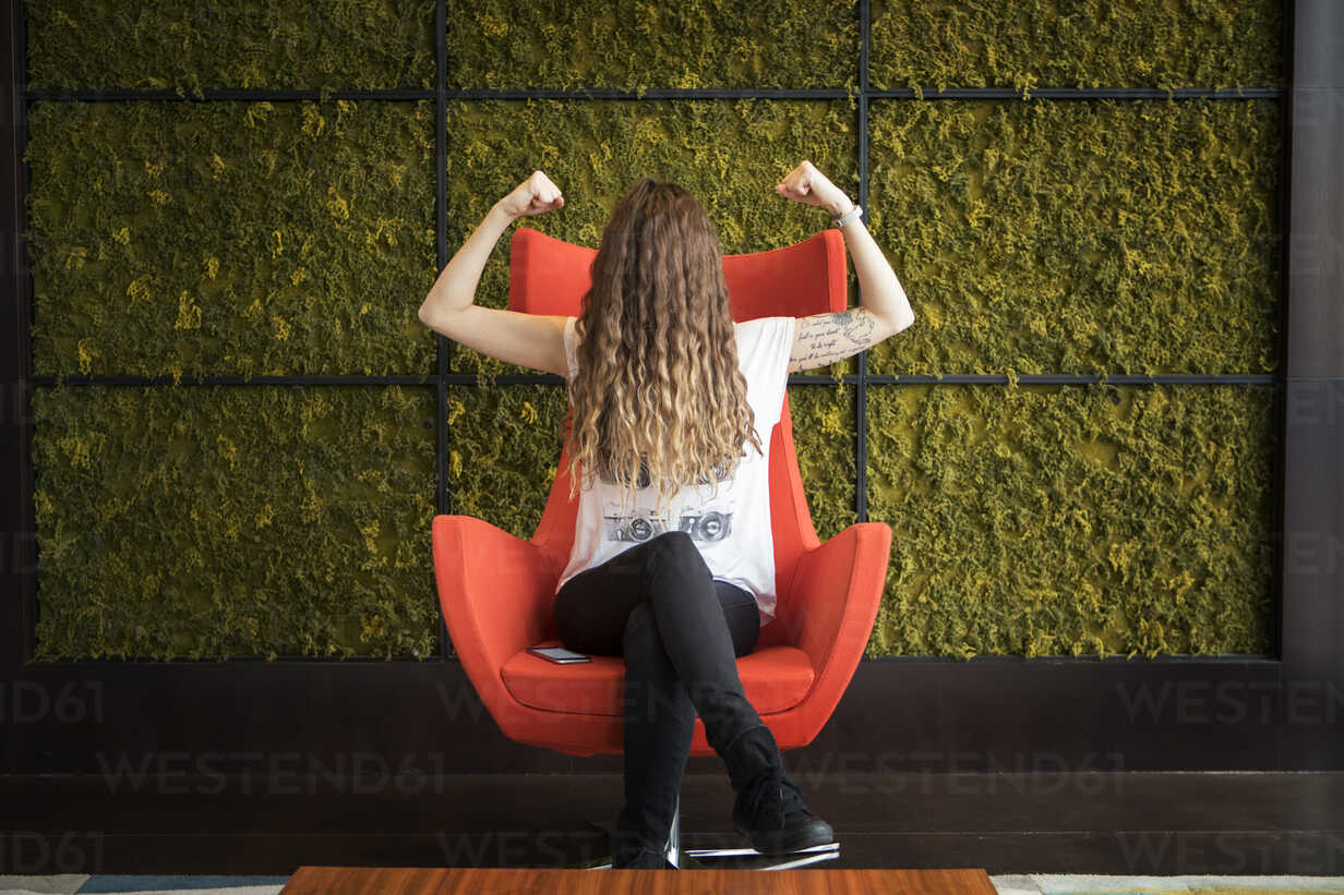 Woman with hair on face flexing muscles while sitting on armchair against wall at home